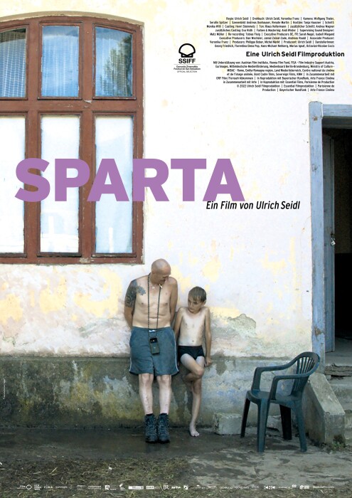 Ewald in shorts and a boy in swimming trunks are standing next to each other leaning against an old house wall. Ewald's head is turned towards the boy in a friendly manner. A large old wooden window, parts of a blue entrance door with a plastic chair next to it can be seen. 