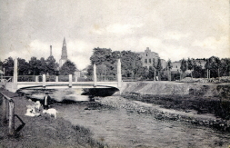  The photo shows a view of the Aa bridge at the Ravarditor from the west shortly after its completion. Passers-by are walking their pets along the unpaved riverbank. The tower of St George's Church can be seen in the background and the Marienlyzeum behind the trees.  