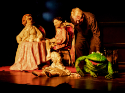 Puppenspiel der Froschkönig Die Königin und der König sitzen in der linken Bildhälfte an einem Tisch. In der rechten Bildhelfte sitzt die Prinzessin vorne auf der Bühne. Der Frosch ist rechts von ihr zu sehen und wird von Matthias Kuchta gespielt