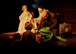 Puppenspiel der Froschkönig - Kinder helfen mit Im Hintergrund sieht man die Königin und den König am Tisch sitzen. Im Vordergrund ist Herr Kuchta mit dem Frosch in der Hand zu sehen. Vor der Bühne stehen ihm gegenüber Kinder.
