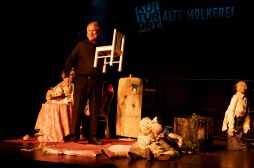 Puppenspiel der Froschkönig - Gesamtbild In der rechten Ecke ist der Diener Heinricht zu sehen. Mittig sitzen die Prinzessin und der Prinz auf dem Boden. Links steht vor dem Tisch mit Königin und König Matthias Kuchta und hält einen weißen Kinderstuhl in der Hand.